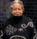 a woman with short salt and pepper hair and light brown skin smiles into the camera. She is wearing a large black turtleneck sweater with spiral designs on it.