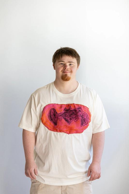 A person wearing a beige t-shirt with a mixed media artwork that resembles a abstract or geometric design in vibrant colors.