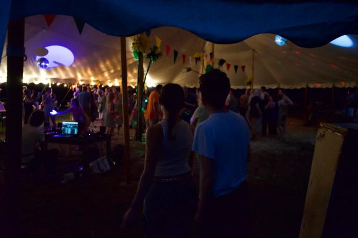 two people stand at the edge of a tent with many people inside it dancing. there are colorful flags and large cellophane flowers, party lights and a hay floor.