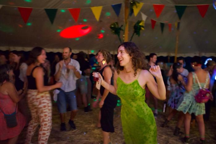 a woman with long curly bron hair dances happily wearing a green dress. in the bakcground are other people dancing in a darkened tent with colorful triangular flags hanging from the ceiling and green party lights shining. theres a red spotlight shining on a discoball and hay on the floor