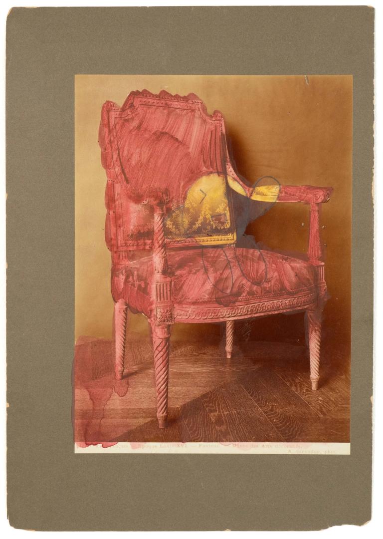 a painting over an old photograph thats mounted on a grey/green board. The photo shows an antique chair on a wooden floor. the painter has painted the chair and floor dark but transparent red, and put a small round cartoonish figure in the chair, painted yellow and with a big smile.