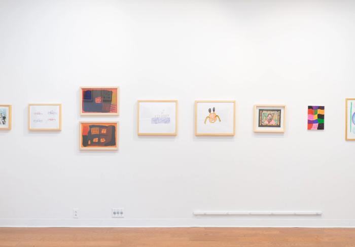 a white gallery wall with nine artworks hanging, all but one framed. there are many different styles