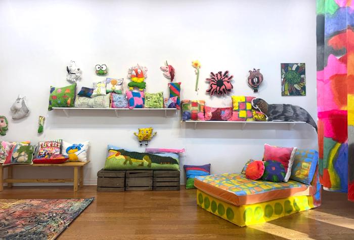 A white walled gallery space, brightly lit, with many colorful obejcts. Theres a painted divan with pillows on it, two shelves with many different pillows of varying shapes and sizes and colors, some hanging on the wall. a corner of a colorful rug is visible and a bench with more pillows.