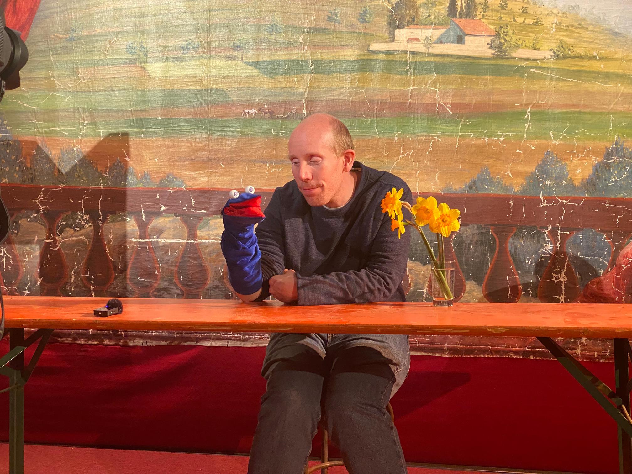 a light skinned man smiles gently down at a blue puppet on his hand in the shape of a bird. the puppet has large white spherical eyes and a bright red mouth. there are daffodils on the orange table he sits at and a painted background of tuscan hills