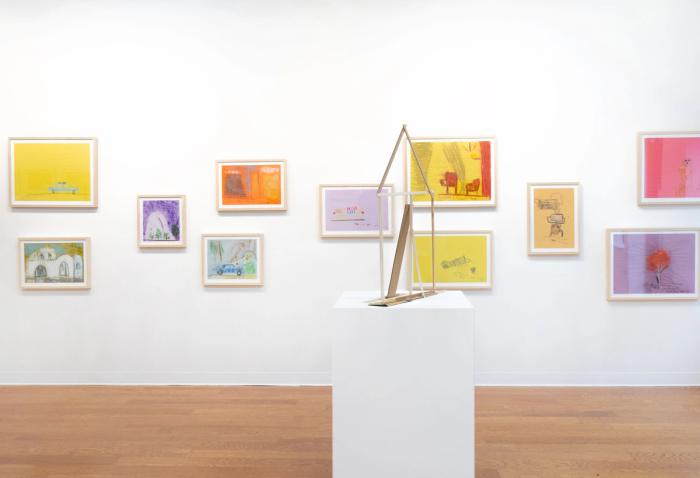 a white gallery wall with many colorful drawings in wooden frames hung in several levels. in front of them is a large pedestal with a thin house shaped sculpture made of pieces of wood 