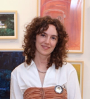a person with pale skin and curly brown hair smiles while standing in front of artworks on a gallery wall