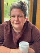 a woman from the chest up smiling towards the camera with a cup of to go coffee in her hand. she has blonde and grey hair put up in a bun with some loose curls on the sides. her eyes are light colored and her skin is light tan.