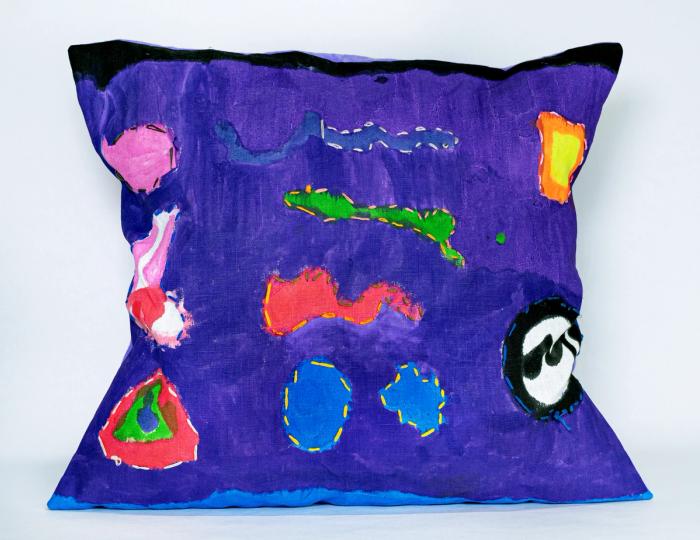 a purple rectangular pillow  with bright abstract symbols all over, many outlined in contrasting stitching
