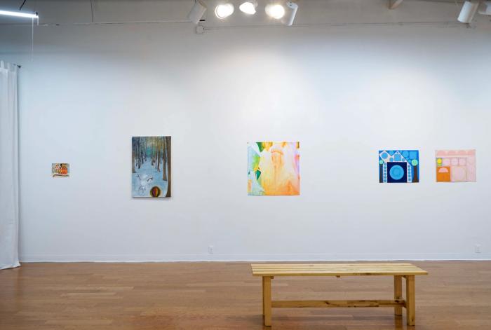 a view of a gallery wall with artworks hanging. colors are bright and the works are diverse, some are abstract and some figurative. the walls are white and there is a wooden bench in the foreground