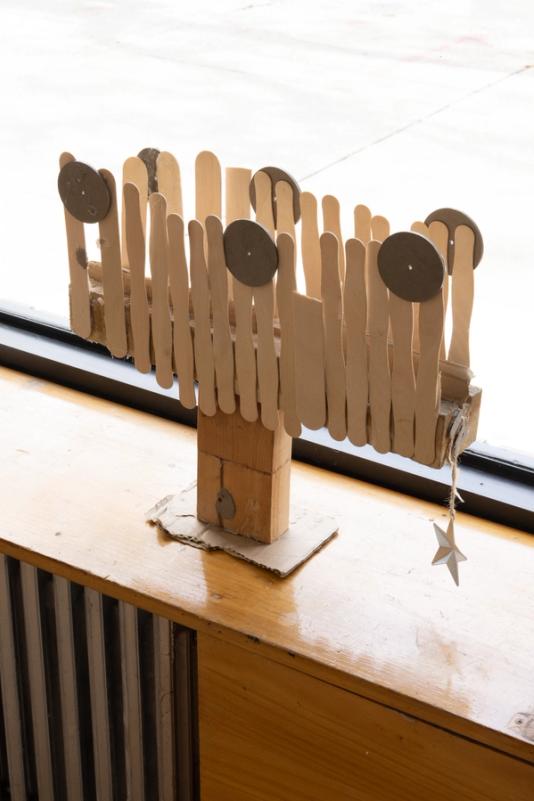overhead view of a sculpture standing on a built in window bench in front of a window looking out on a street on a bright day. the sculpure stands about a foot tall and two wide and has a wooden base, clean popsicle sticks upright in a loose fence-like formation and three evenly spaced circular forms on the top. hanging off the right side is a silver star. 