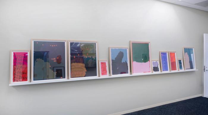 a long shelf with many framed artworks on it. all are abstract paintings with a few colors taking predominance and another color accenting in small hash marks here and there. some have text running the length of them