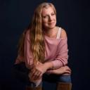 a woman with long slightly wavy blond hair parted and hanging down to the left side of her face sits in a dark space looking at the camera. shes smiling gently and wearing a pink sweater, ripped jeans, and tall light brown boots.