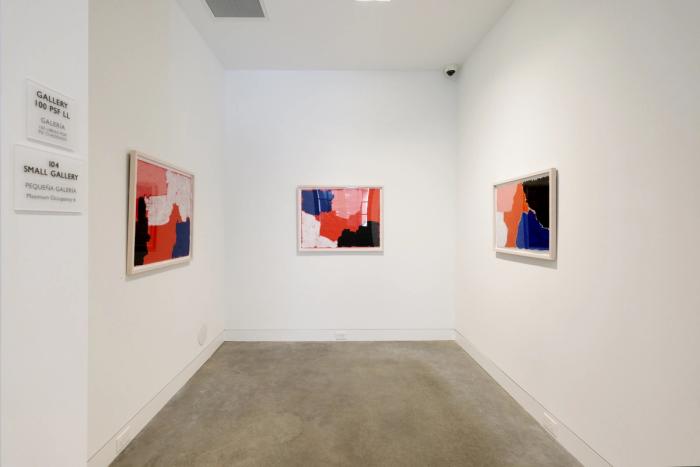 a corner of a white walled gallery room with three walls visible and three abstract paintings in frames hanging