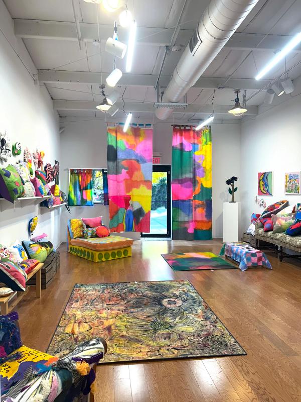 A white gallery space with lots of colorful objects in it. Theres a rug in the foreground with loose paintings of flowers, shelves with many pillows in many shapes and colors, and in the back two very tall curtains hanging with large color-fields painted on them.