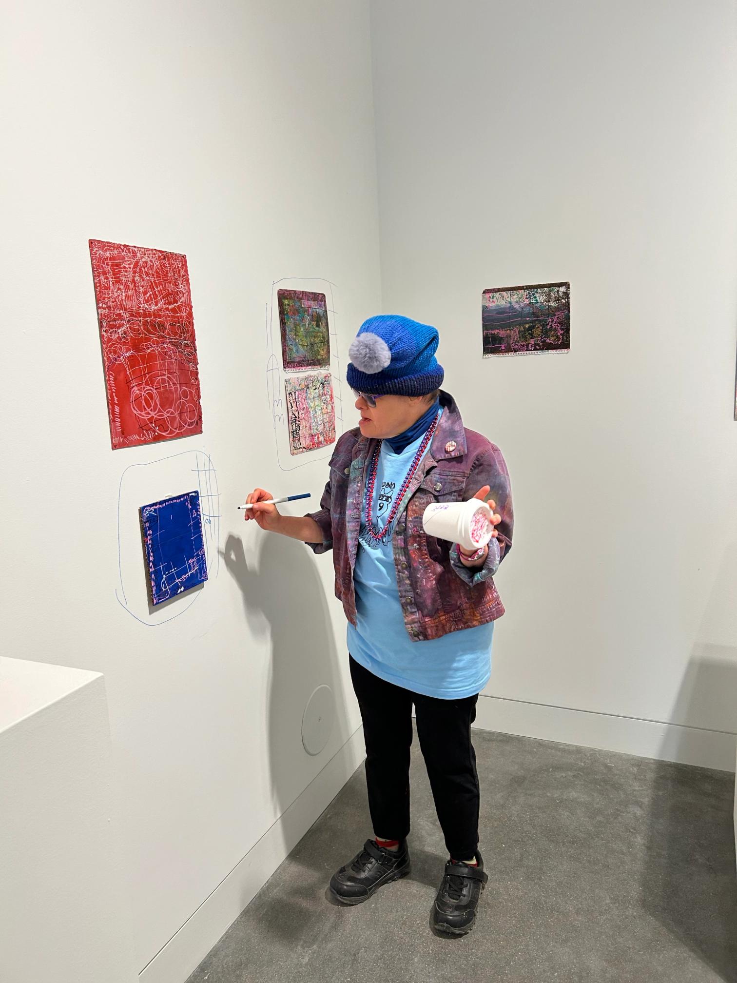 a woman with light skin stands in front of several artworks installed in a white walled gallery space. she holds a coffee cup with some marks on it and is poised to draw onto the wall around the artworks.