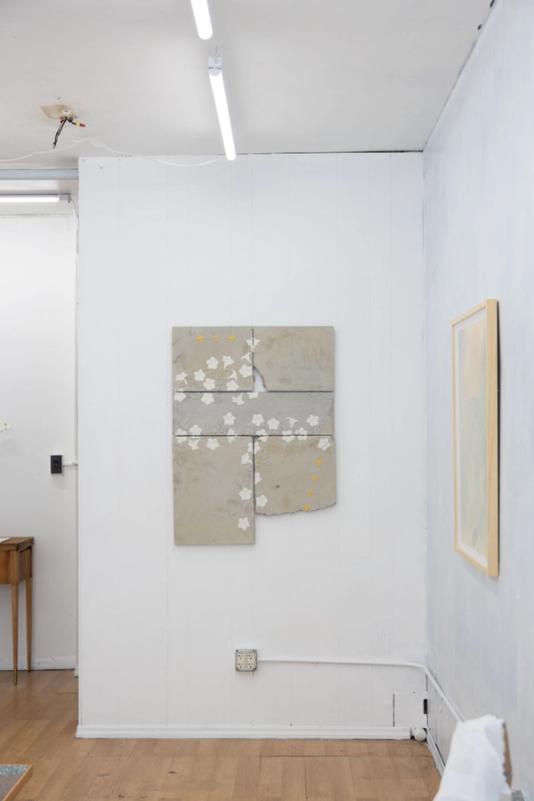a well lit gallery space with bead board walls and vinyl wood flooring. in center of the image is a large work made of cast concrete thats done in 5 sections. there are white flowers strewn across but mostly around the dges where the pieces meet. several yellow bees fly through.
