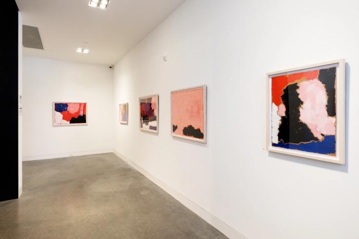 a corner of a white walled gallery room with two walls visible and five abstract paintings in frames hanging