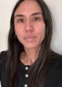 a woman with long black hair looks into the camera. she has tan skin and dark eye color and angular features. she wears a black shirt with white buttons and a medium smallgold hoop is visible in her ear.