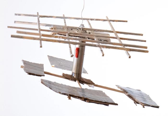 a silver sculpture hanging from a wire made of sticks crisscrossing, conencted by a silver flashlight to a set of crossed sticks with pieces of rectangular cardboard on the end.