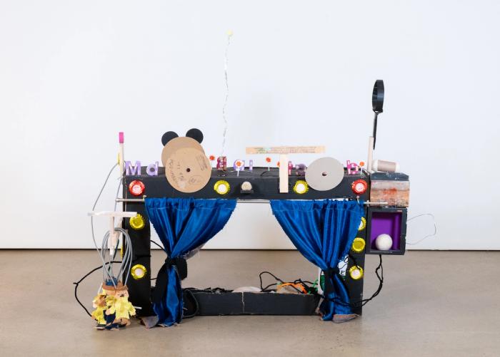 A mixed media sculpture resembling a television set and/or DJ turntables, featuring knobs, letters, circular discs, and blue velvet curtains.