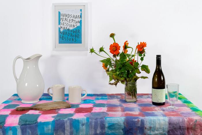 an install photograph of the Living Room exhibition, featuring a custom-made blue, pink, and red checkerboard tablecloth by artist Eric Spencer. A ceramic pitcher, two mugs, a cutting board, a vase with orange flowers, a wine bottle, and a glass are arranged on the tablecloth. Behind the table, a framed painting by Michael Angelo Mangino is displayed. It is a blue and white abstraction with overlaid handwritten text that reads, "handshake perception sessions make over wardrobe lessons."