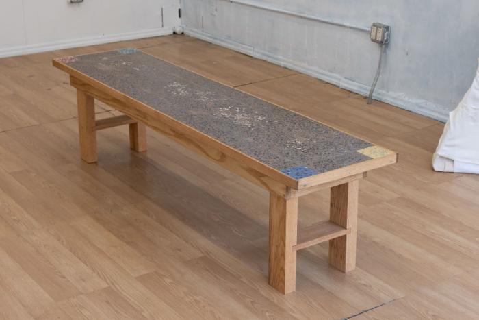 a wooden framed bench with a very intricate pebbly cement-like top section thats inset into the wooden frame. most of the top is dark gray brown with each corner containing a small square of a different color, red, blue/green, yellow, dark blue. 