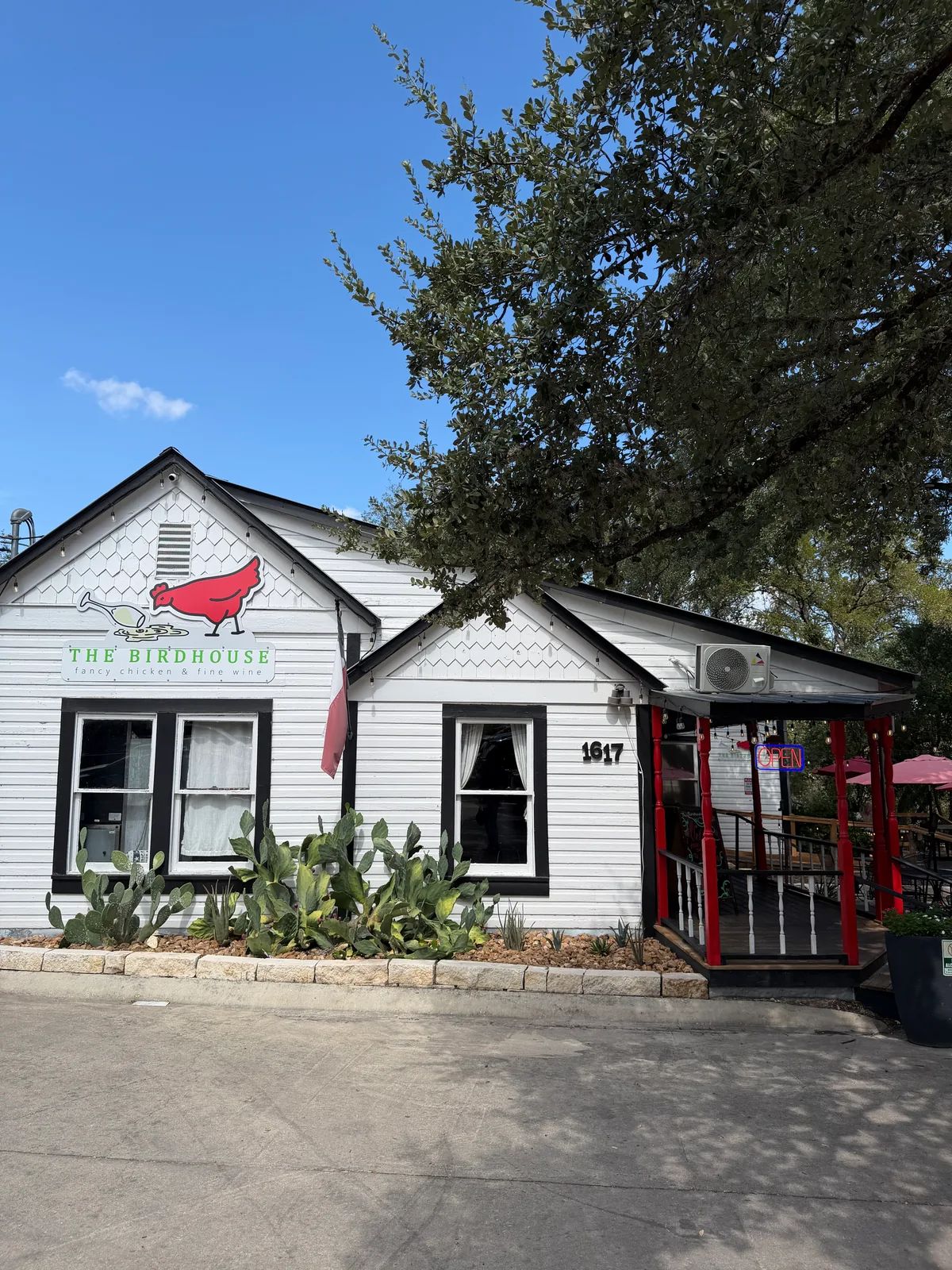The front of The Birdhouse, a white cottage with a red bird on its sign, cactus planted along the front and red posts on the porch, under a large tree