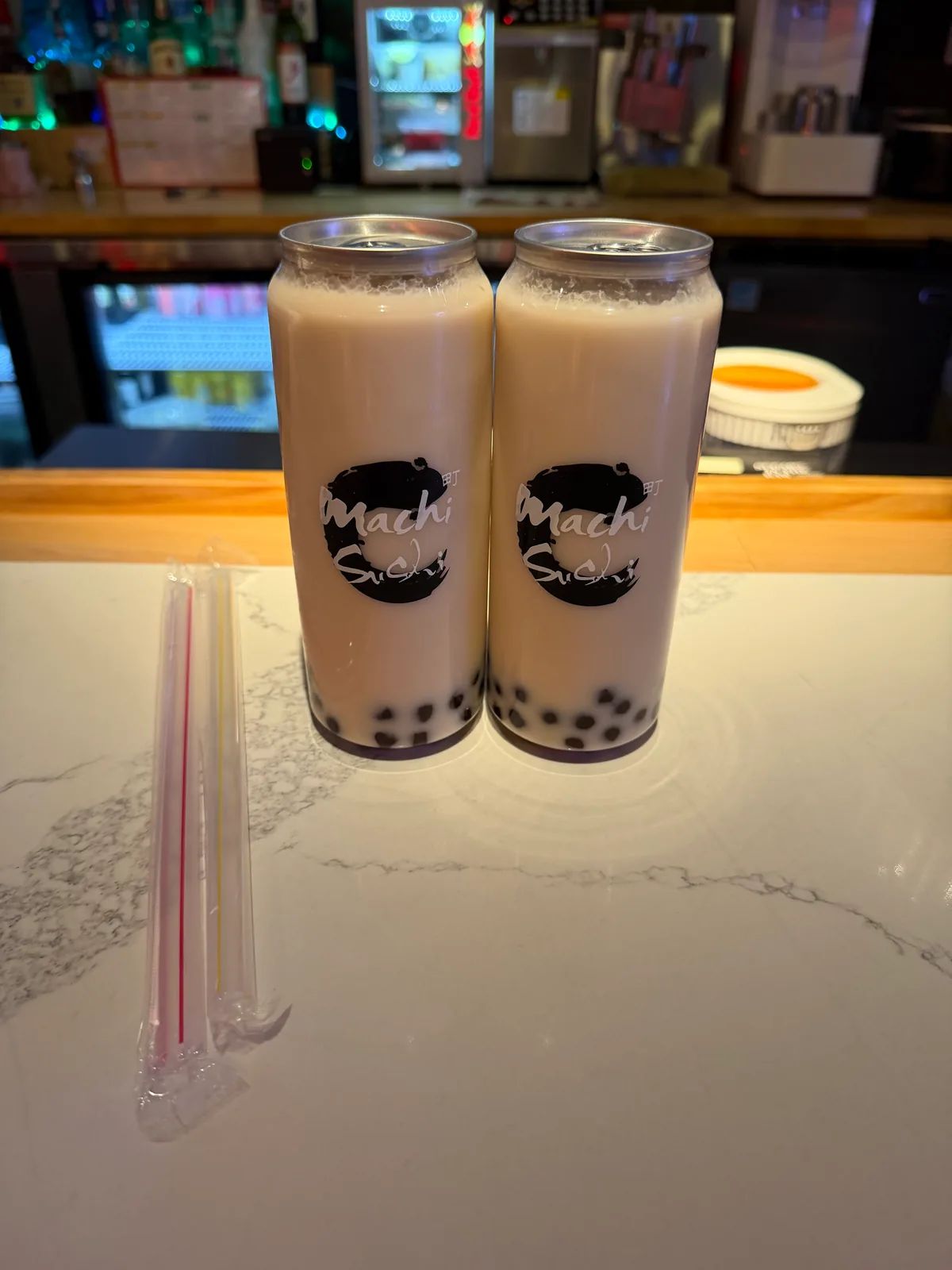 Two cans of milk tea with boba, printed with the Machi Sushi logo, next to two wrapped straws on a marble counter