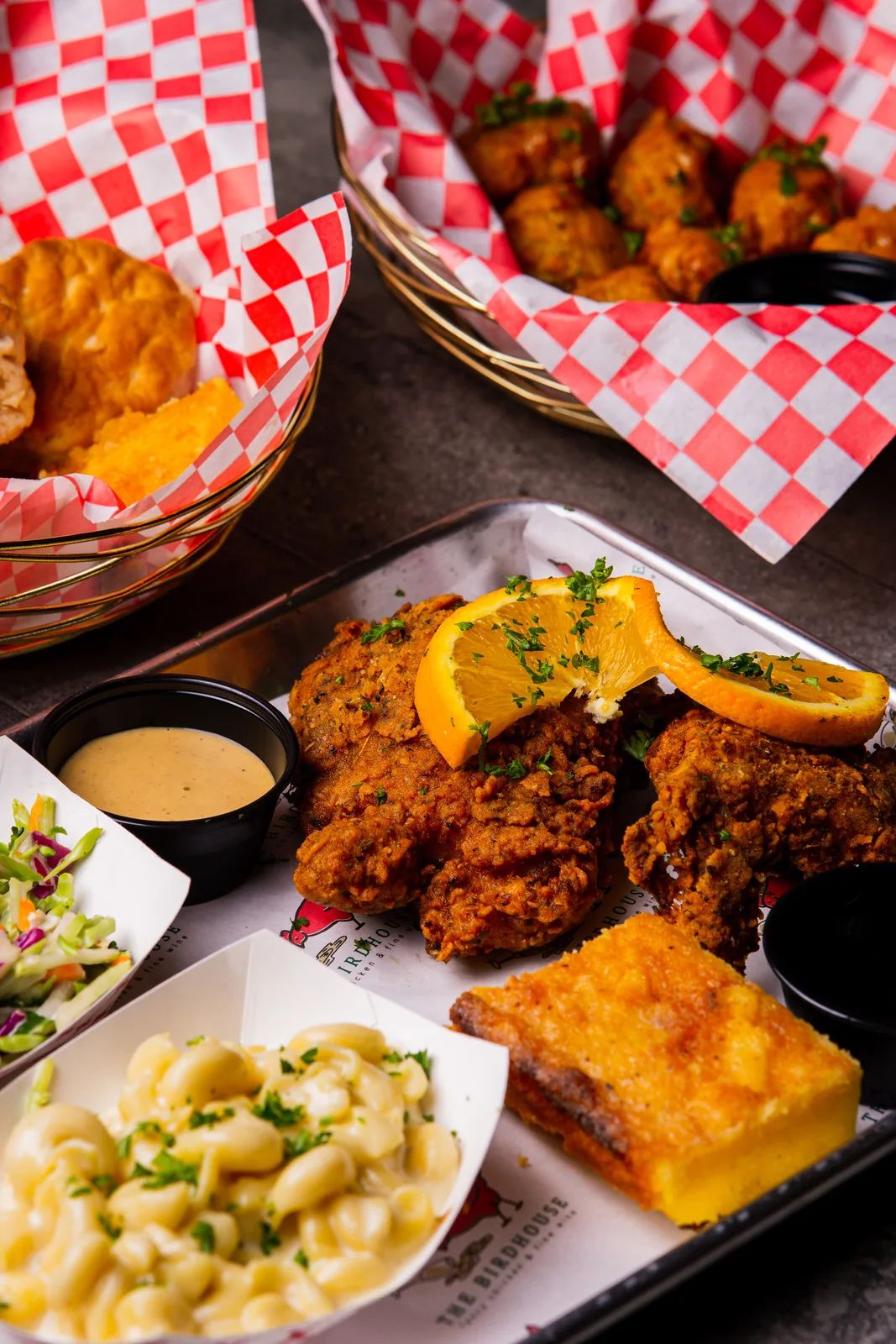 A fried chicken plate topped with an orange slice, with mac and cheese, slaw, a square of cornbread and a dipping sauce, and checkered baskets behind