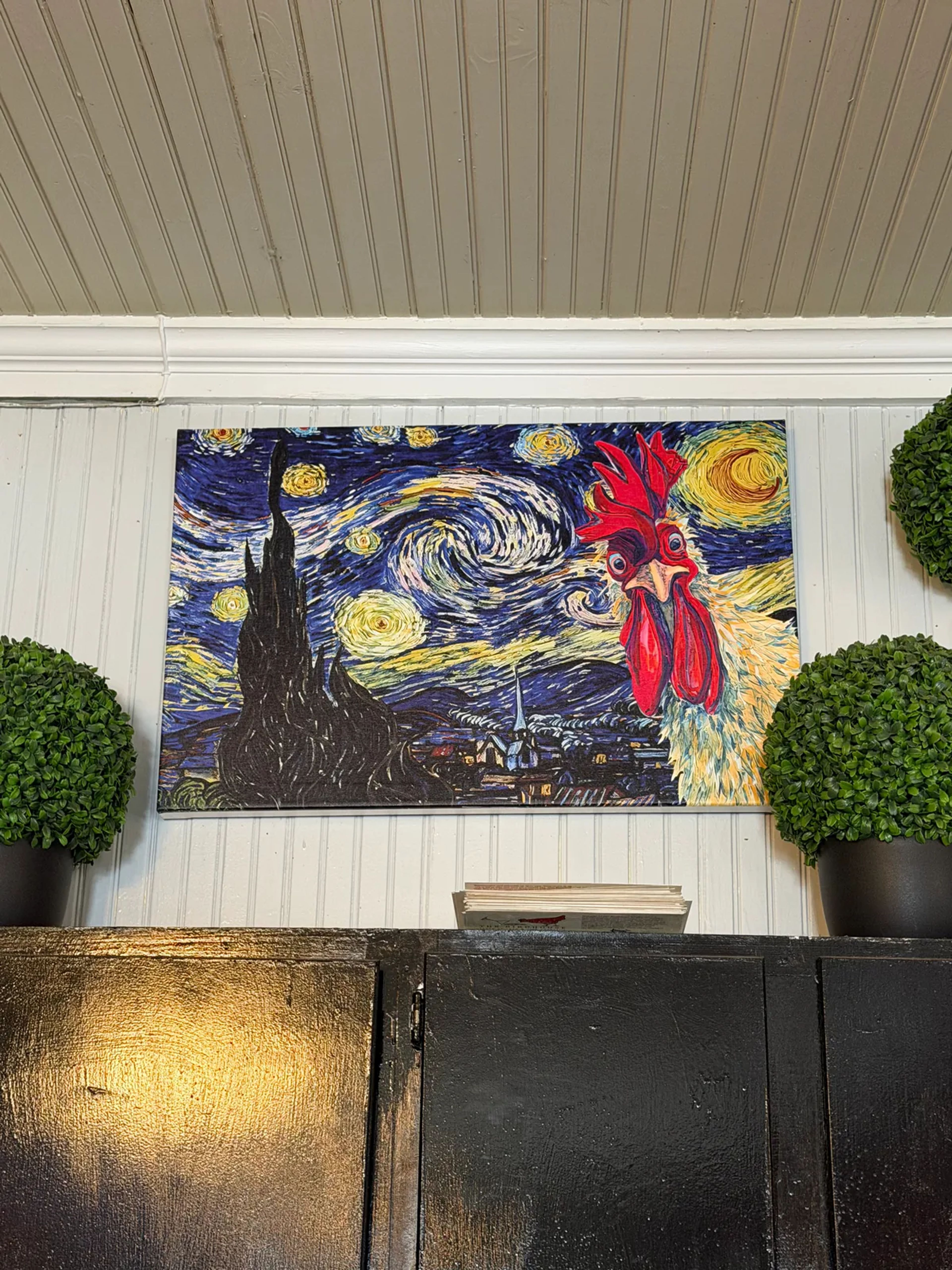 A Starry Night print with a red rooster looking in from the right, hung between two topiary balls above a black cabinet