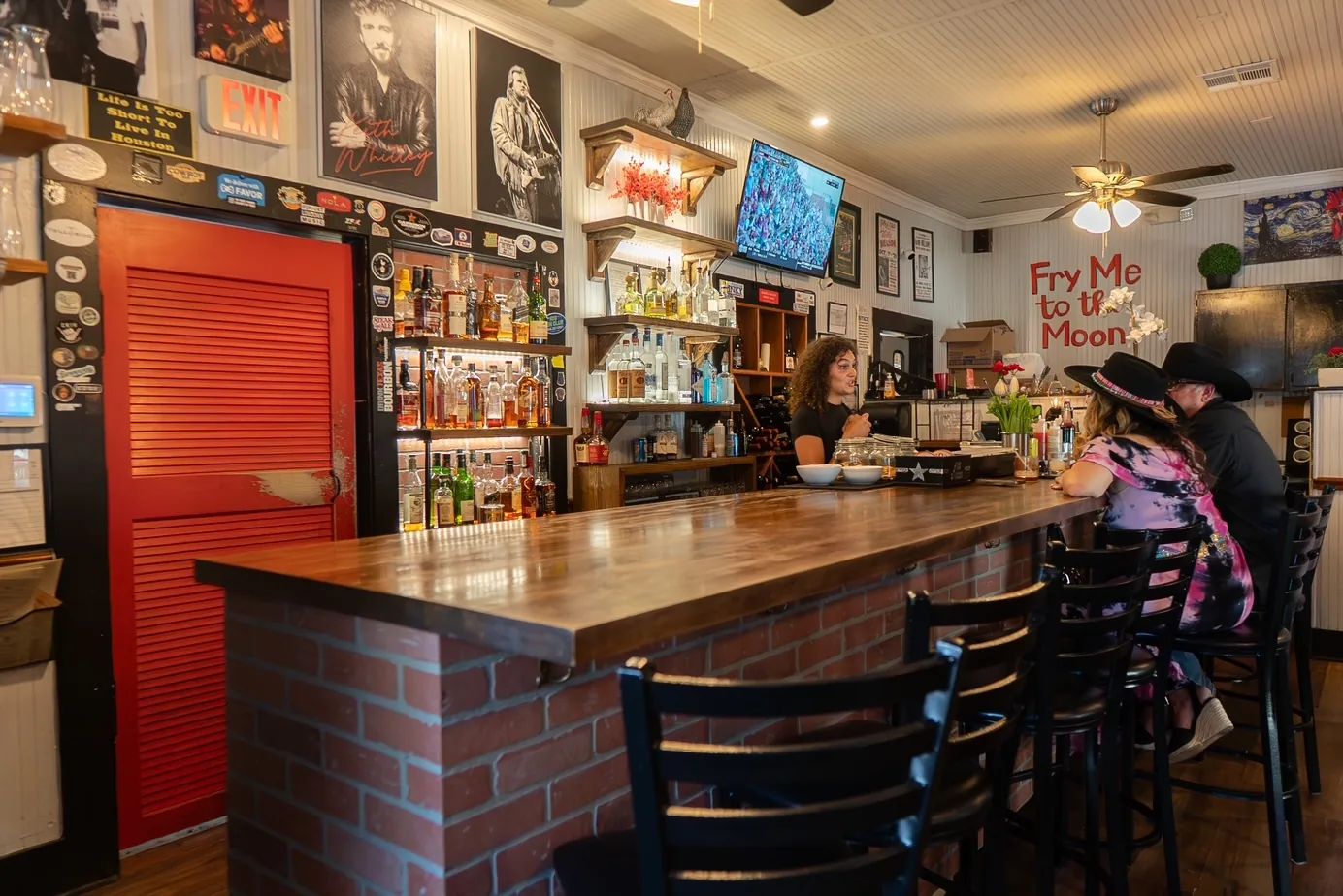 The Birdhouse bar, with a brick front and black stools, a bartender talking with guests in cowboy hats, music posters above and Fry Me to the Moon in red letters on the wall