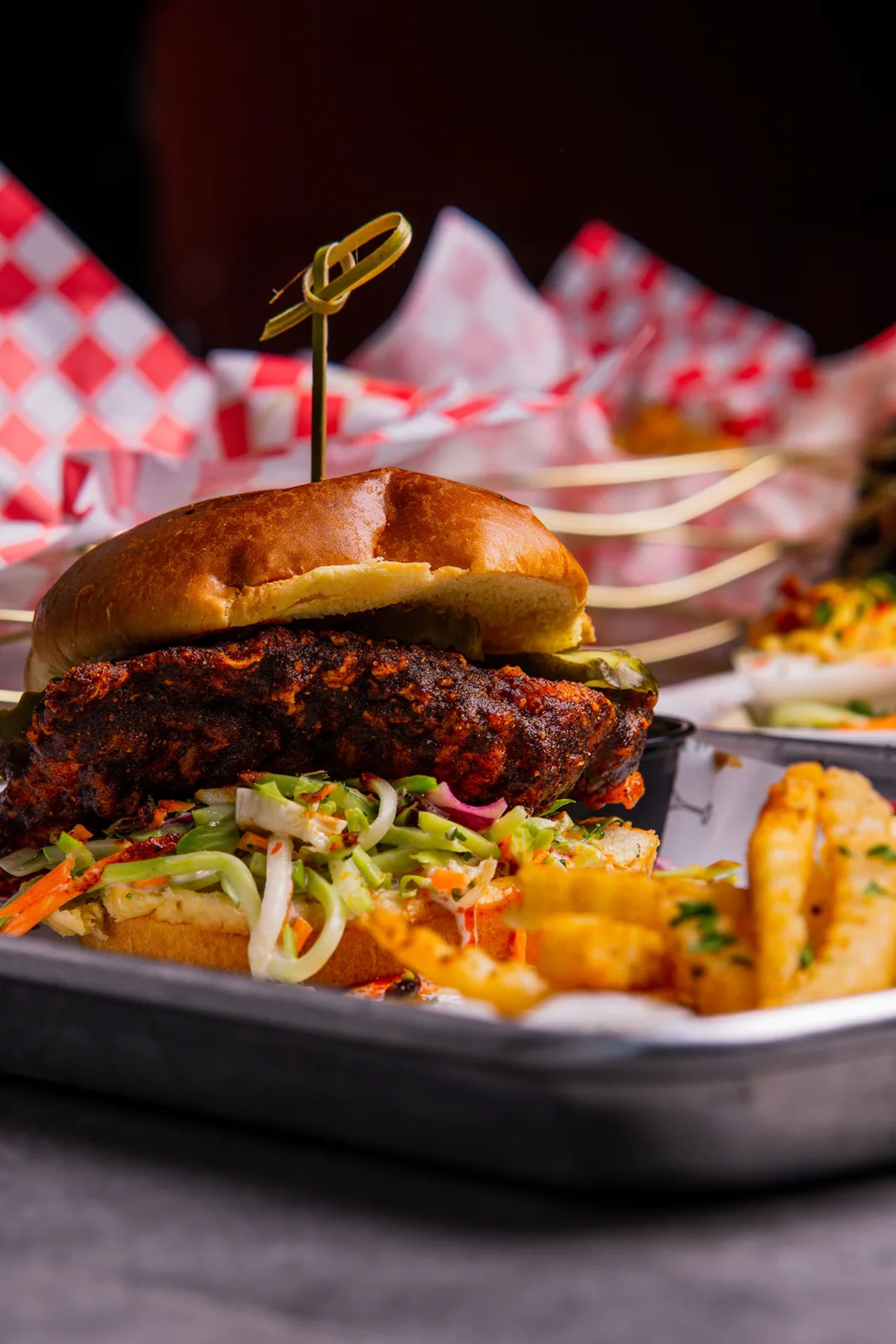 A fried chicken sandwich with slaw and a pickle on a toasted bun, with crinkle fries on a metal tray