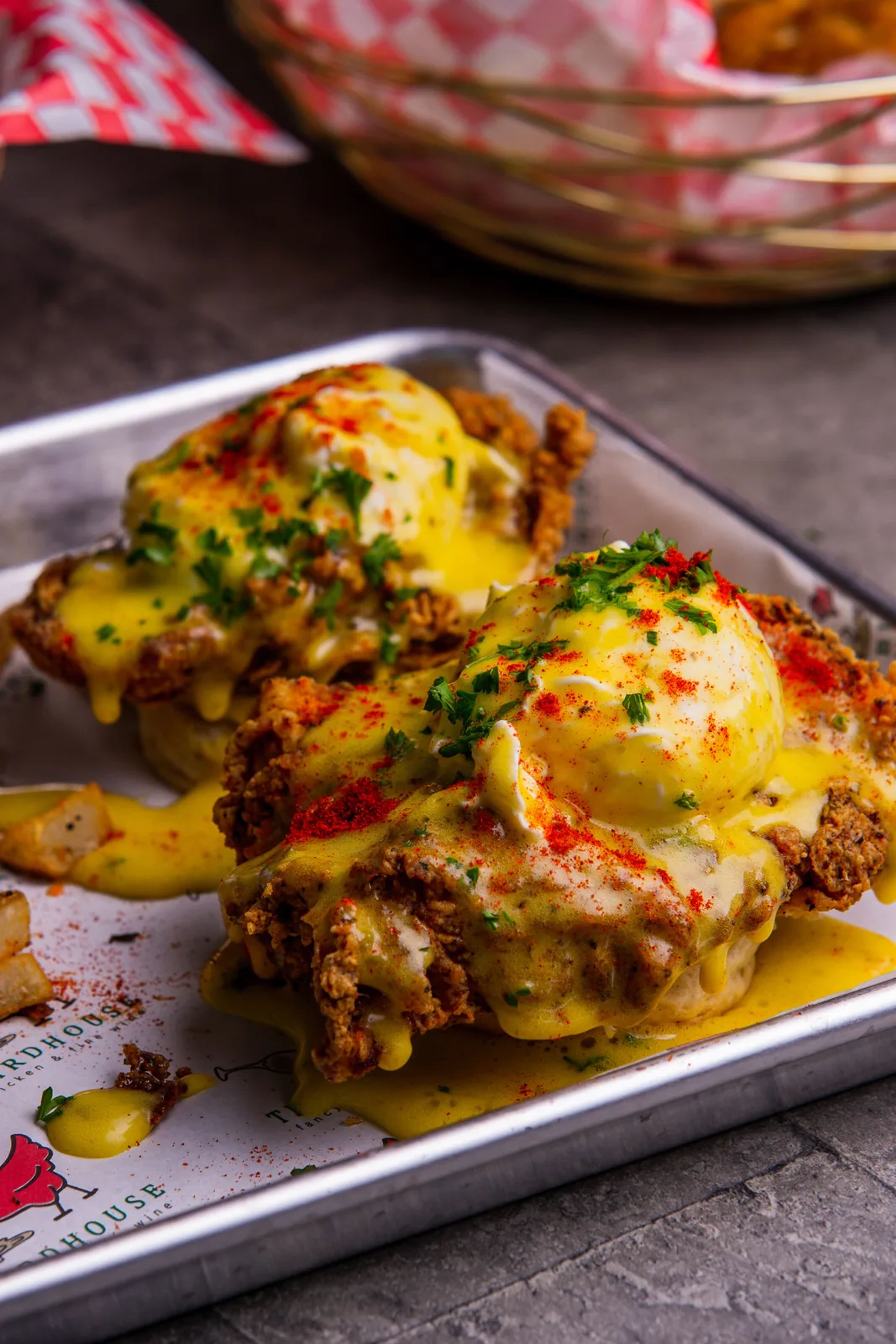 Two pieces of fried chicken topped with poached eggs, hollandaise, paprika and parsley on a lined metal tray, with fried potatoes, and a checkered basket behind