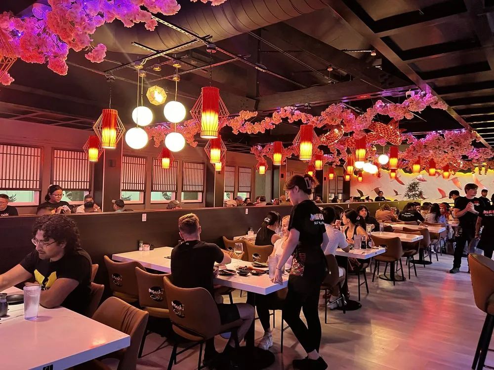 The Machi Sushi dining room full of diners under red lanterns and pink blossom garlands, with a server standing at a table