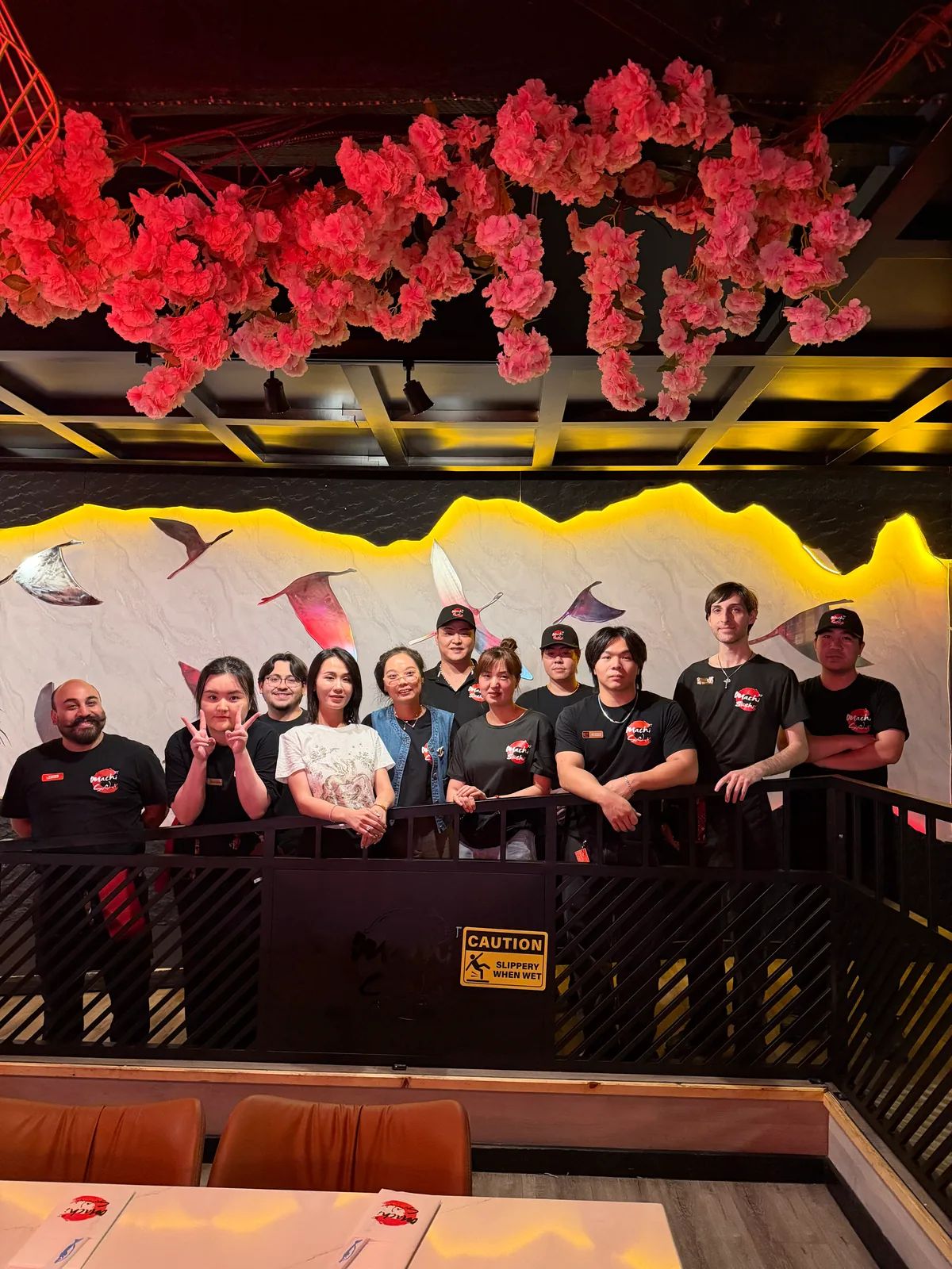 The Machi Sushi team lined up along a railing under pink blossoms, in front of a wall of bird sculptures