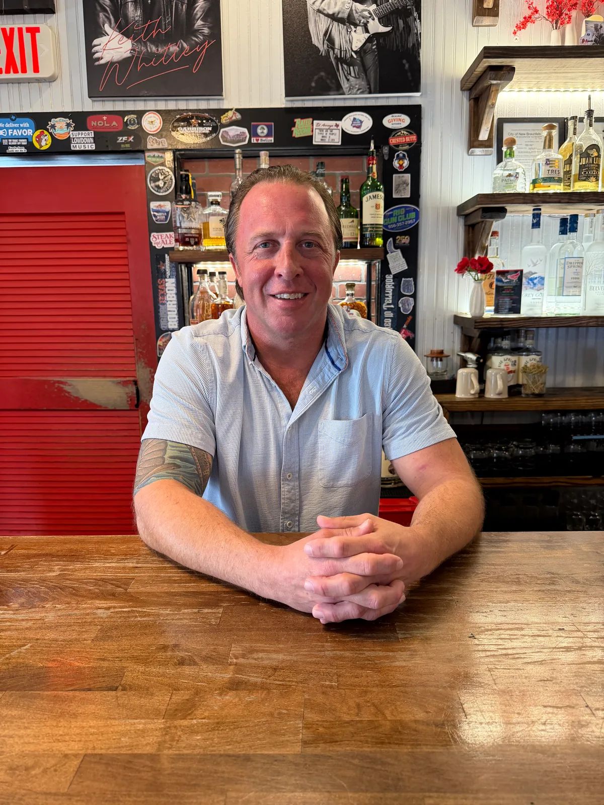 Ross Wilkinson, owner of The Birdhouse, smiling with his hands folded on the wooden bar, a red louvered door and shelves of bottles behind him