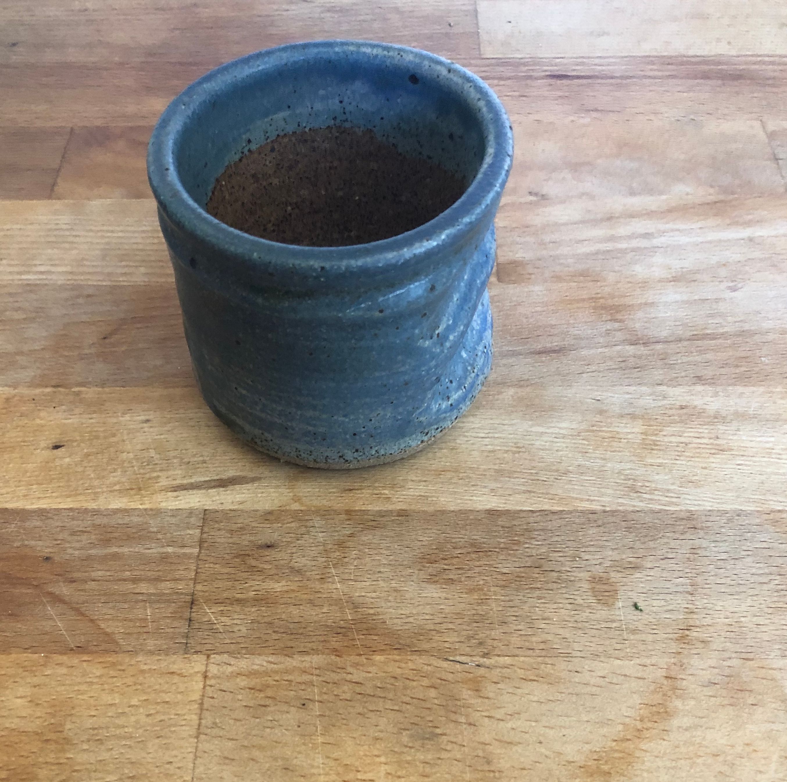 a small dark and muted blue ceramic glazed cup with no handles on top of a light wooden surface. On the side of the cup there are three indentations which create a space for someone to place their fingers into. The view of the cup is slightly from a bird's eye view and shows the inside of the cup which has a natural dark brown color and clear gaze. 