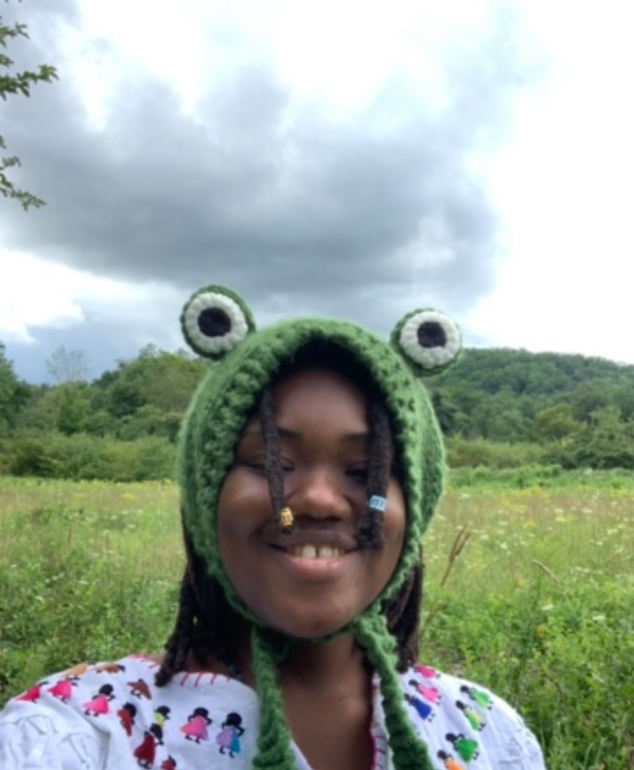 Rowan is standing on a field in front of a rolling green hill. The foliage and grasses behind them are made of greens, yellows, and browns. Rowan is wearing a white shirt with embroidered figures decorating the neckline of the shirt - the shirt also has red stitching around the collar. Rowan has a green frog knit hat on top of their head - it has two eyes and ties at the bottom. The sky behind Rowan appears slightly cloudy and gray. 