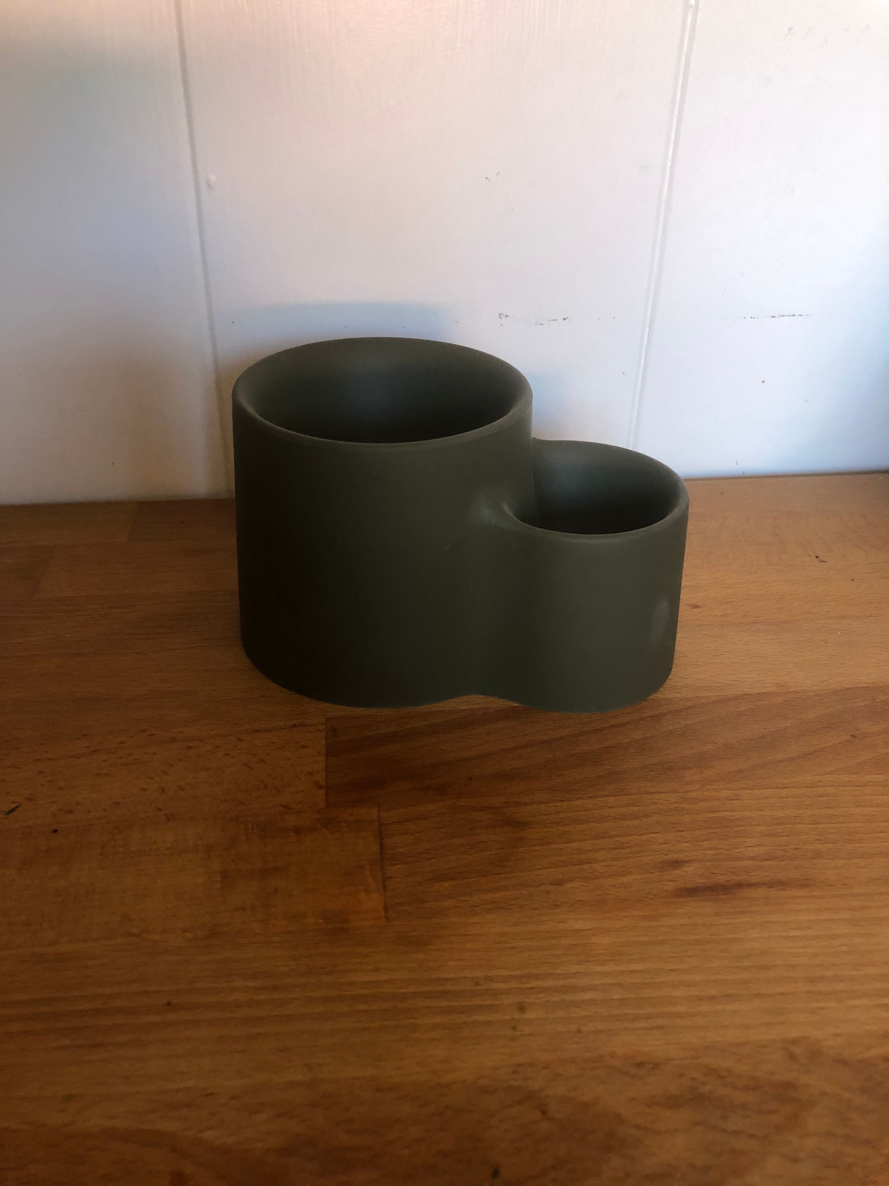 A silicone forest green vessel on top of a wood surface in front of a white wall. The vessel is made up of two bowl-like cylinder shapes connected to one another. The first cylinder is larger in diameter and taller in height then the second one which it is connected with. 