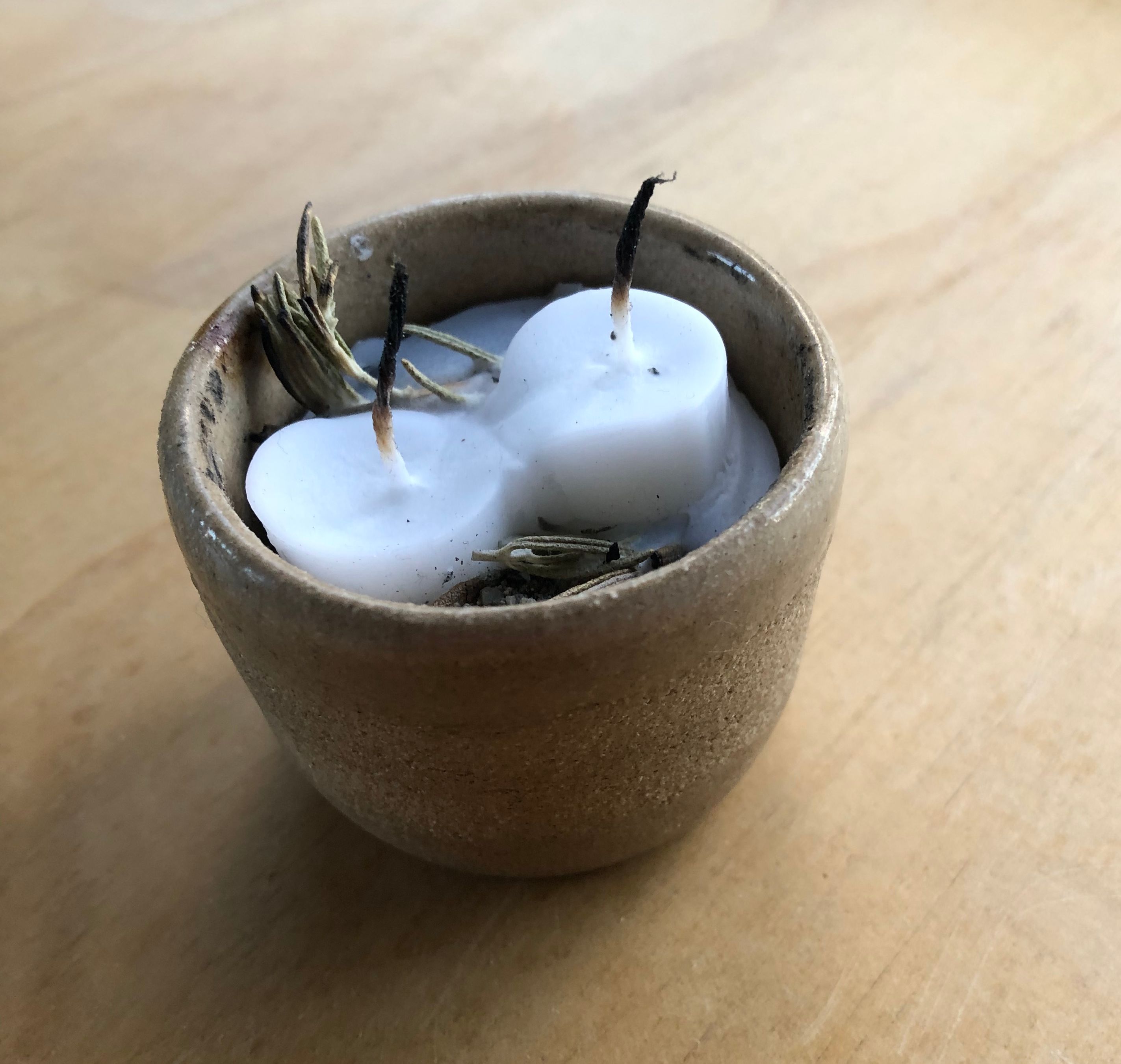 A close up photo of a small beige sand colored bowl sitting atop a ply wood counter top. Inside of the vessel there are two melted white shabbat candles. There is dried rosemary partially submerged with the melted wax. 