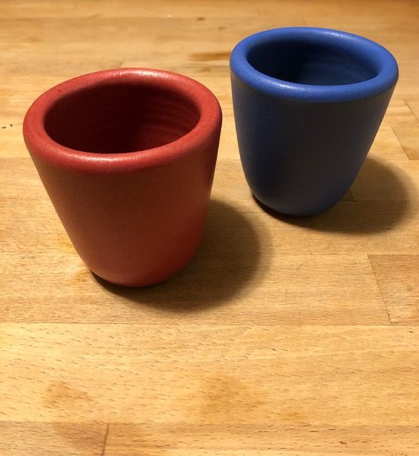 Two ceramic cups sitting on top of a light wooden counter. The cup on the viewer's left is red and the cup on the viewer's right is royal blue. The two cups are casting a small circular shadow and the red cup is slightly in front of the blue cup.