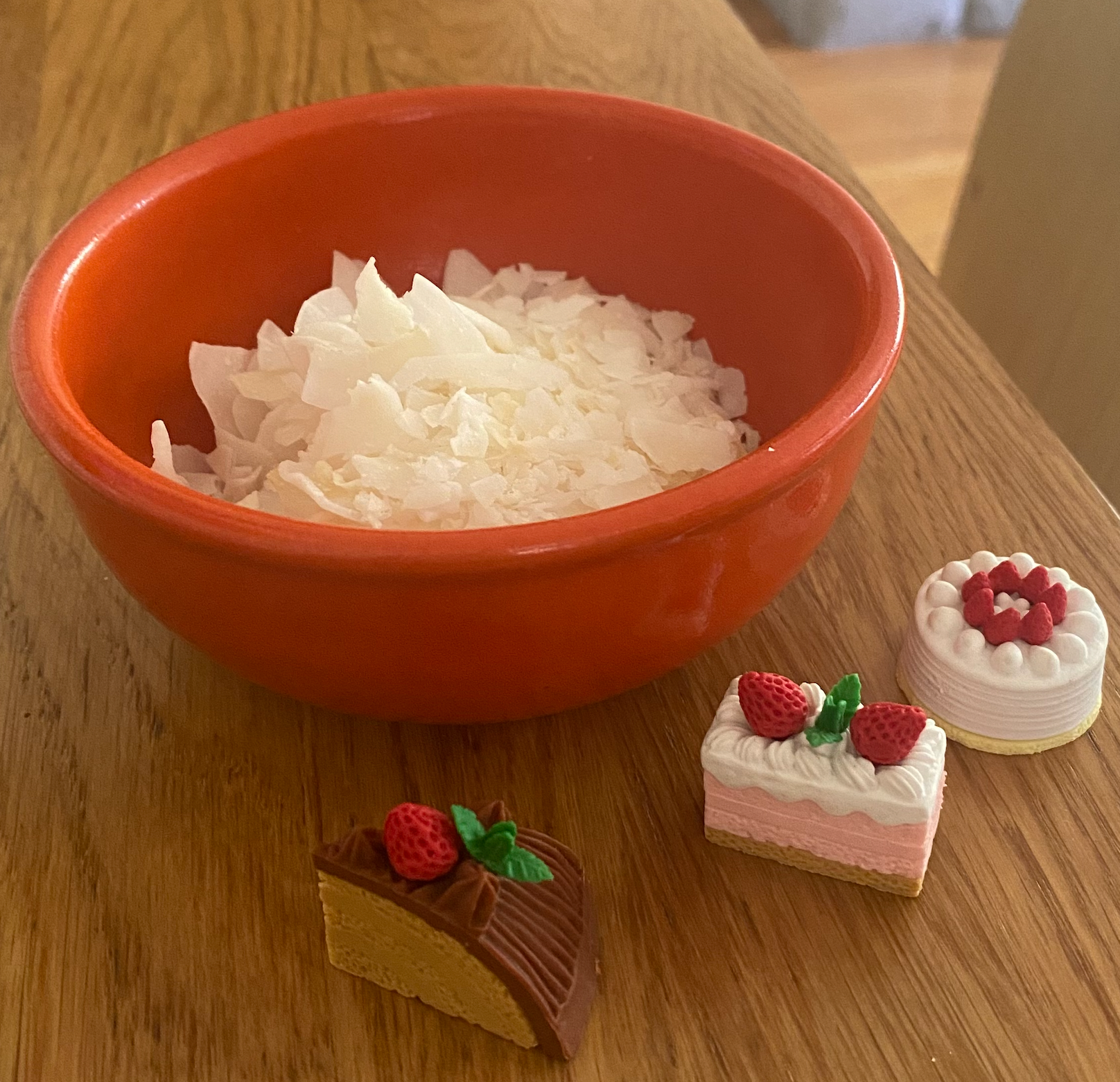 A small orange bowl filled with coconut flakes. In front of the bowl there are three miniature erasers all shaped like different kinds of cake. The first cake has a beige cake with chocolate frosting and a small red strawberry. The second has a strawberry light pink filling with white frosting and two strawberries. The third is a small full vanilla cake with white frosting and red strawberries on the top in a circle 