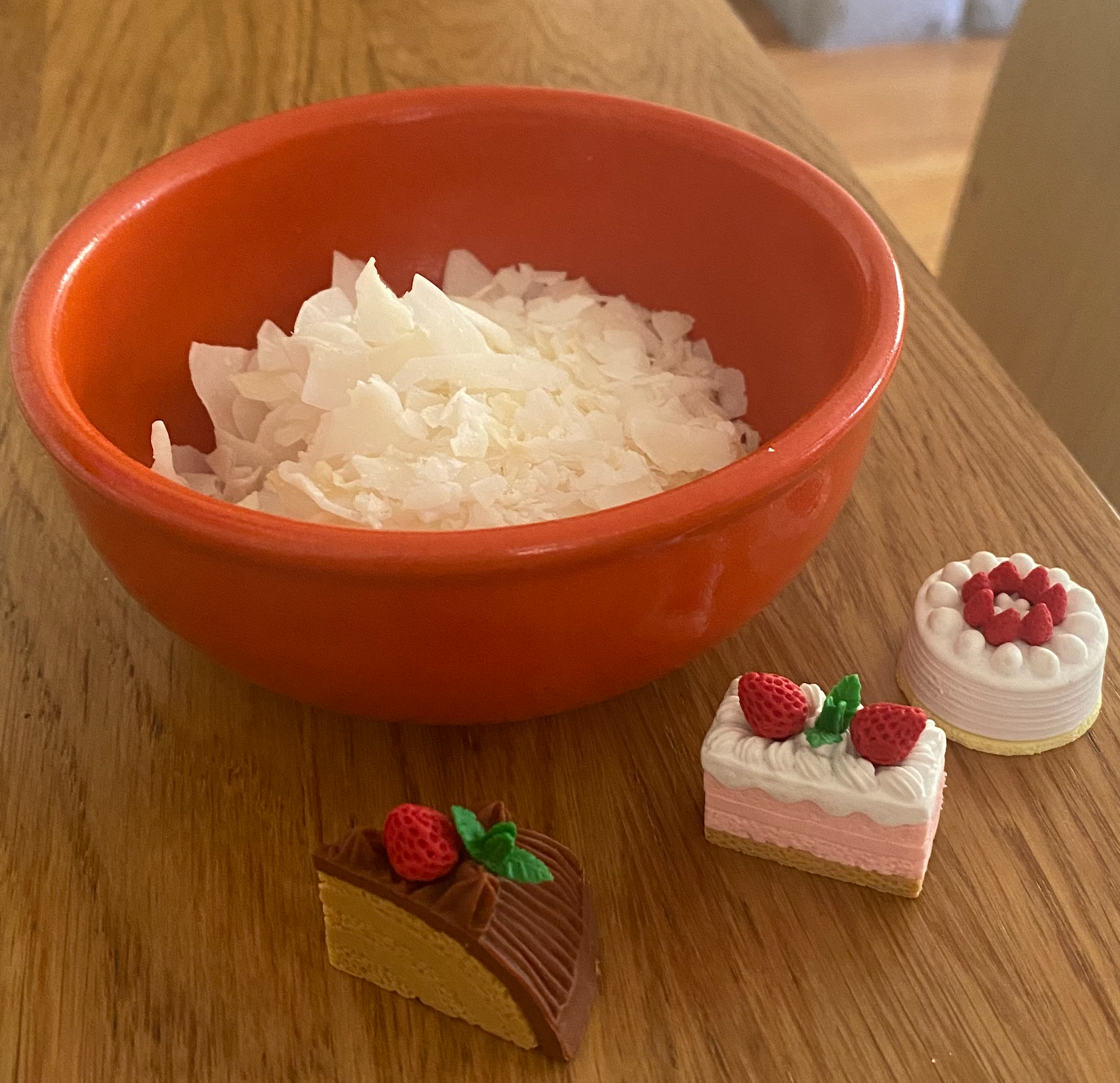 A small orange bowl filled with coconut flakes. In front of the bowl there are three miniature erasers all shaped like different kinds of cake. The first cake has a beige cake with chocolate frosting and a small red strawberry. The second has a strawberry light pink filling with white frosting and two strawberries. The third is a small full vanilla cake with white frosting and red strawberries on the top in a circle