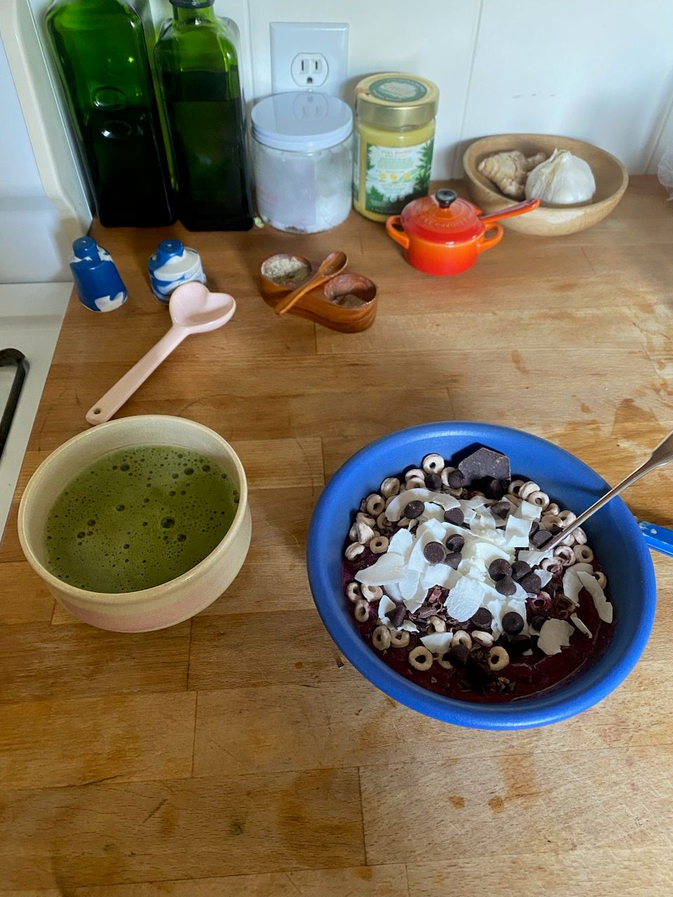 two ceramic vessels sitting on top of an oak wooden kitchen counter. On the left there is a porcelain chawan filled with bubbling matcha green tea. To the right there is a matte blue ceramic bowl filled with a dark purple smoothie topped with coconut shreds, cheeriors, dark chocolate chips and a metal spoon. Behind the vessels there is a blue and white ceramic salt and pepper shaker, a glossy ceramic pink heart spoon, and containers filled with olive oil, ghee, coconut oil, and garlic/ginger. There is also a small orange and red la creuset mini pot.  