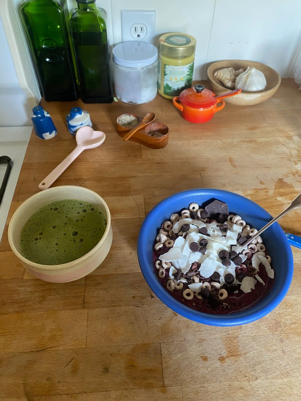 two ceramic vessels sitting on top of an oak wooden kitchen counter. On the left there is a porcelain chawan filled with bubbling matcha green tea. To the right there is a matte blue ceramic bowl filled with a dark purple smoothie topped with coconut shreds, cheeriors, dark chocolate chips and a metal spoon. Behind the vessels there is a blue and white ceramic salt and pepper shaker, a glossy ceramic pink heart spoon, and containers filled with olive oil, ghee, coconut oil, and garlic/ginger. There is also a small orange and red la creuset mini pot.