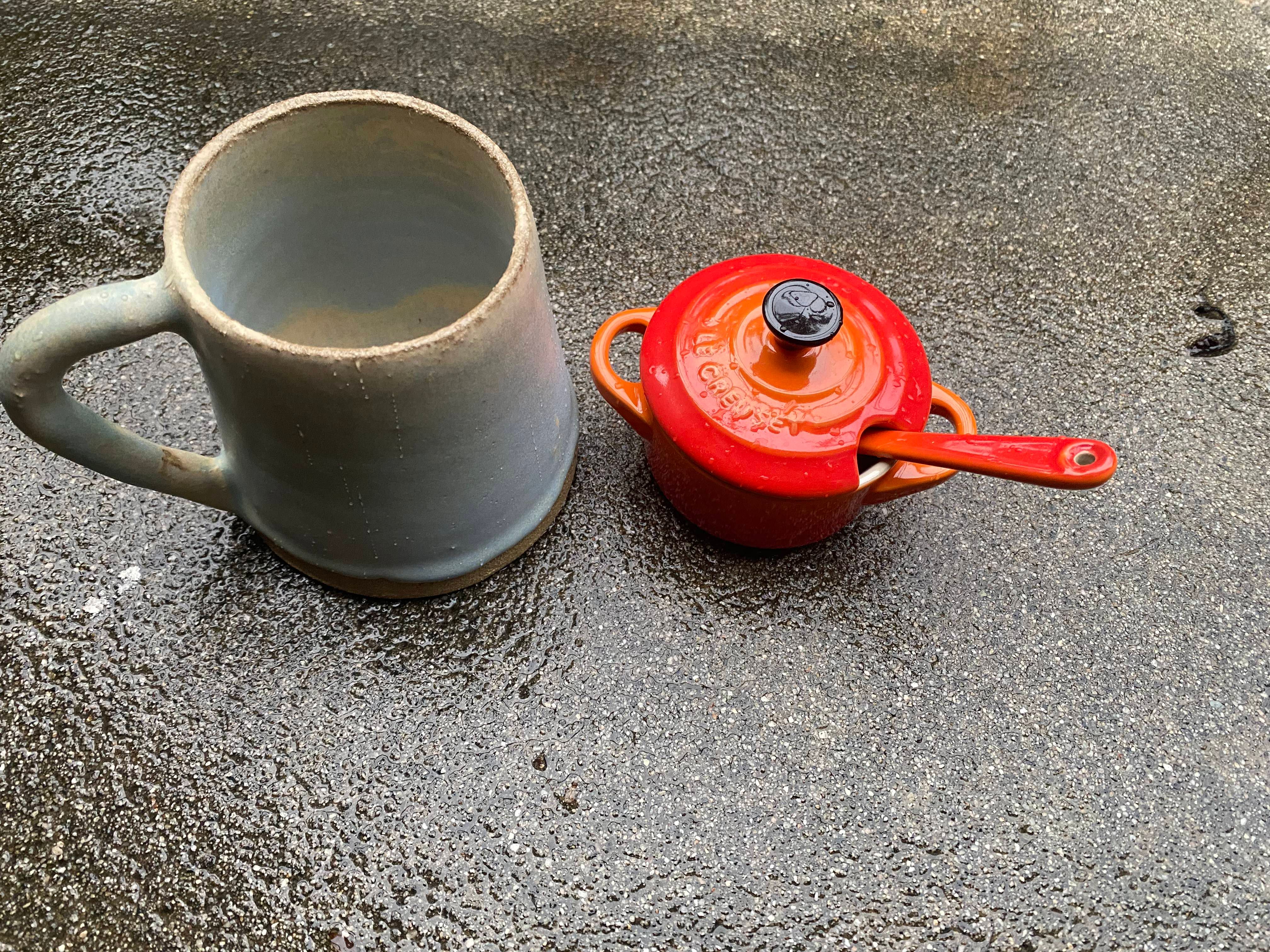 a light blue and off white ceramic mug with a handle to the left of a mini orange and red La Crueset pot sitting atop a wet and dewey cement floor. The vessels are wet with rain.