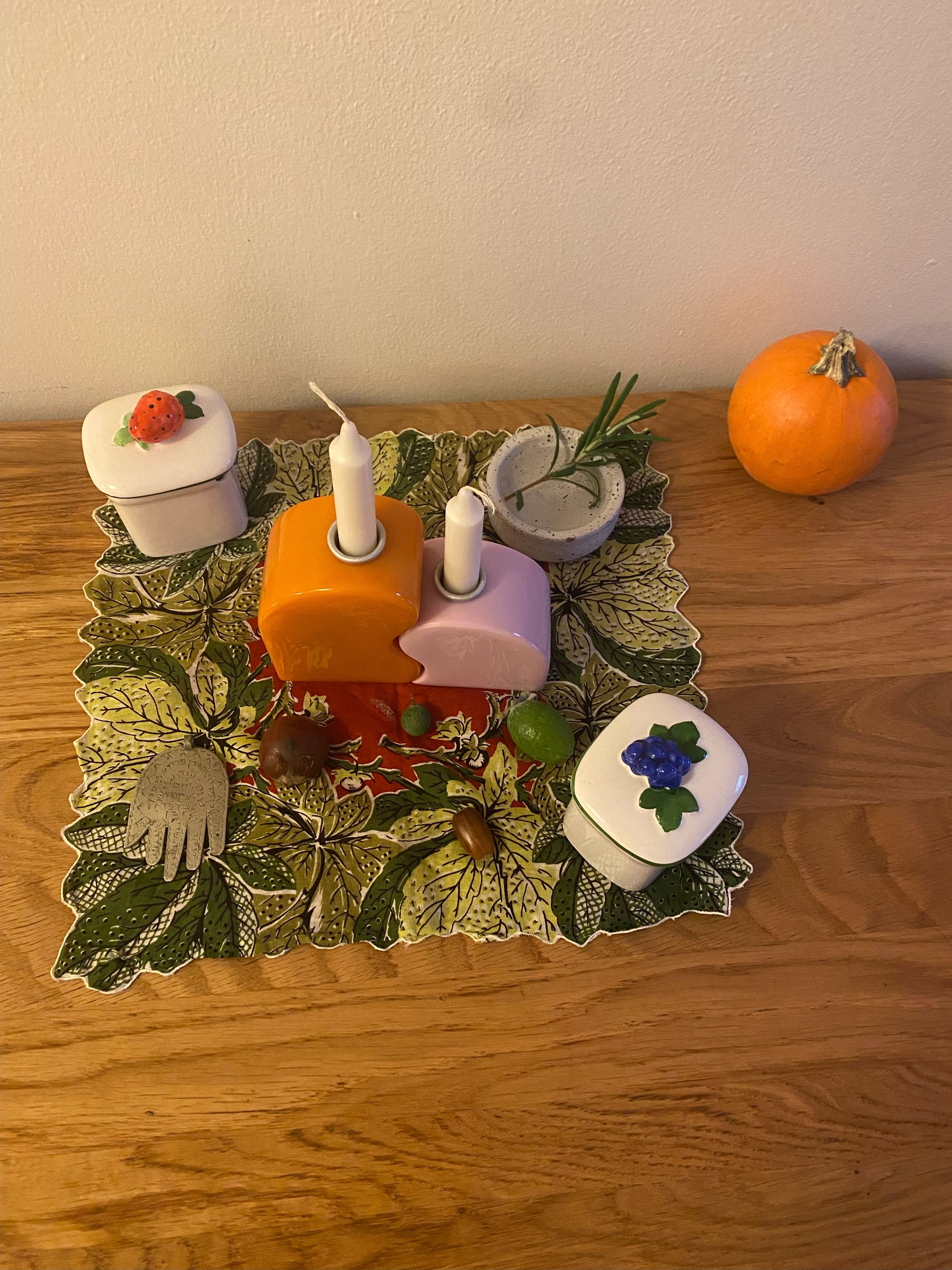 a square textile with green leaves and a red center on top of a Oak wood table. On top of the textile which has rippled edges, there are two white porcelain vessels one with a red porcelain strawberry on top and one with dark blue porcelain blueberries on top. There are two candle holders which fit together like wavey puzzle pieces - one side is orange, the other light lavender/pink. There is a small white ceramic bowl with water and rosemary as well, which sits to the left of a small orange pumpkin. There is also a metal chamsa amulet, 1 acorn, 1 piece of da zao (chinese red jujube date), and two small passion fruits.