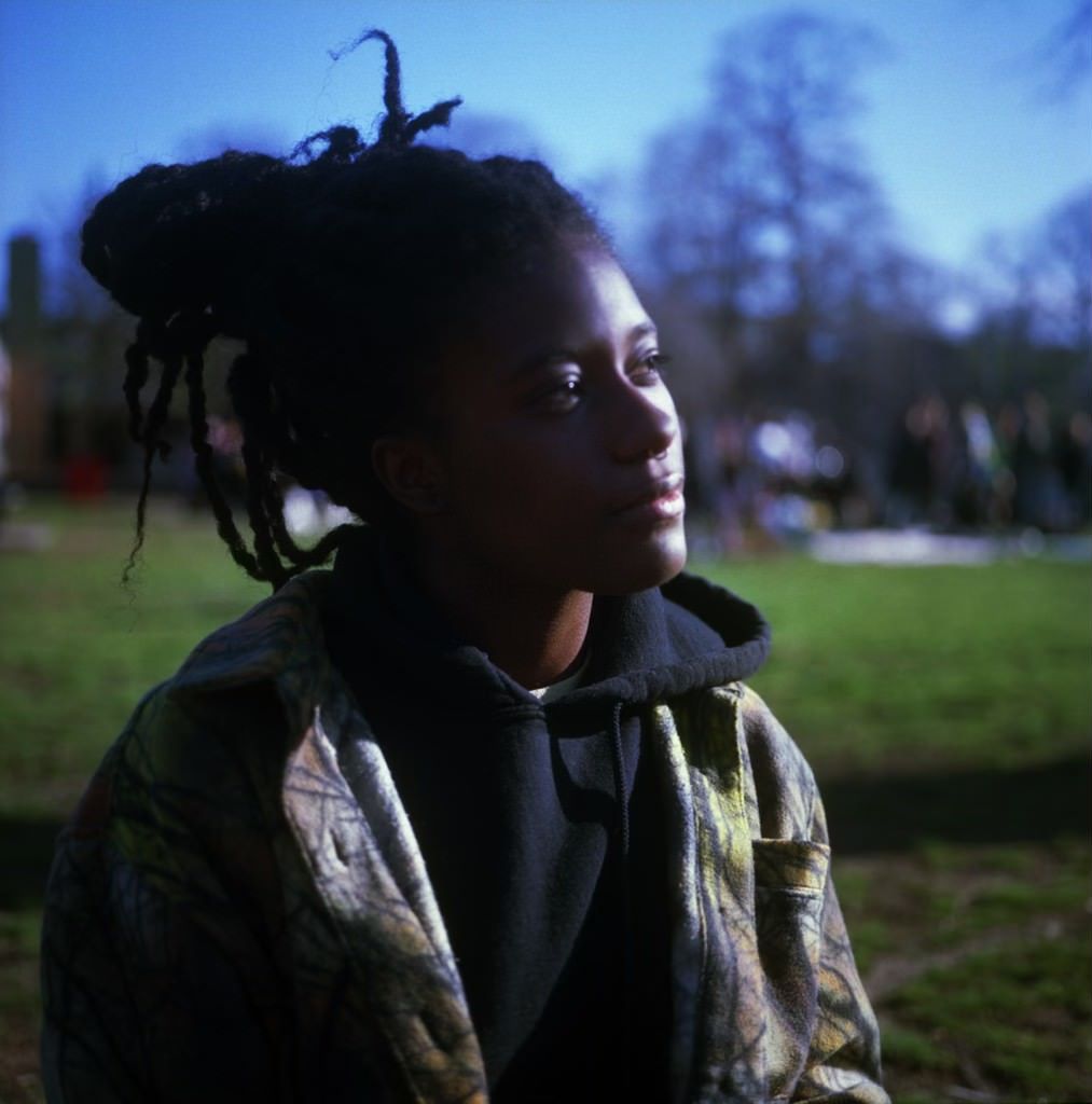 a photo of Cy X sitting on a field of vibrant green grass. Cy is wearing a black hooded sweatshirt under a green, black, yellow, and gray jacket. They are looking into the distance 