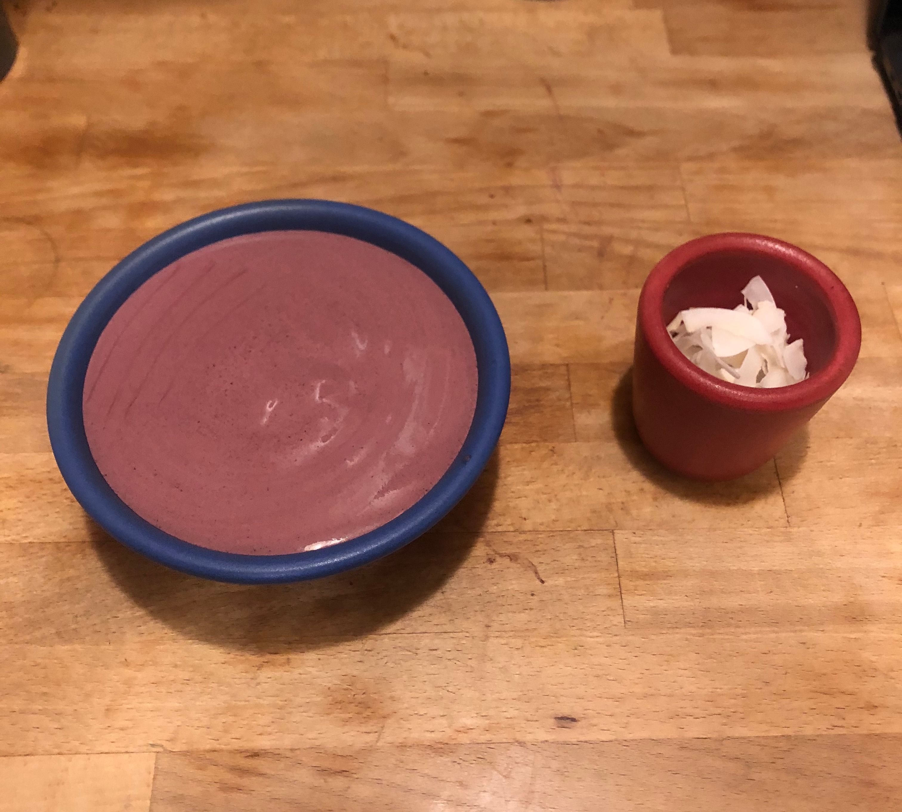 A blue ceramic bowl to the left of a red ceramic cup. The blue bowl is holding a dark purple viscous smoothie and the red cup is holding white coconut shreds. Both vessels are sitting on top of a light wood surface.