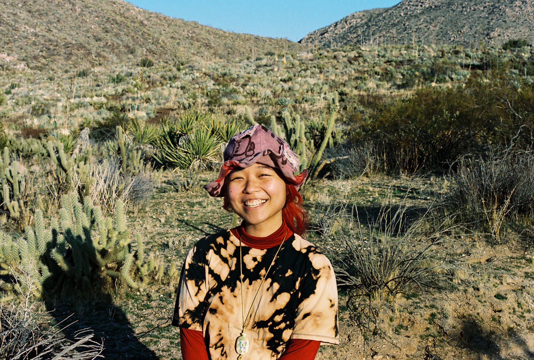 A photo of Rino Kodama standing in front of a vast valley made up of green and brown foliage and cactus. Rino is wearing a red longsleeve shirt with a brown and beige tie dye shirt on top, as well as a pink and red and purple bucket hat. Their hair is red and is pulled back. They are smiling.